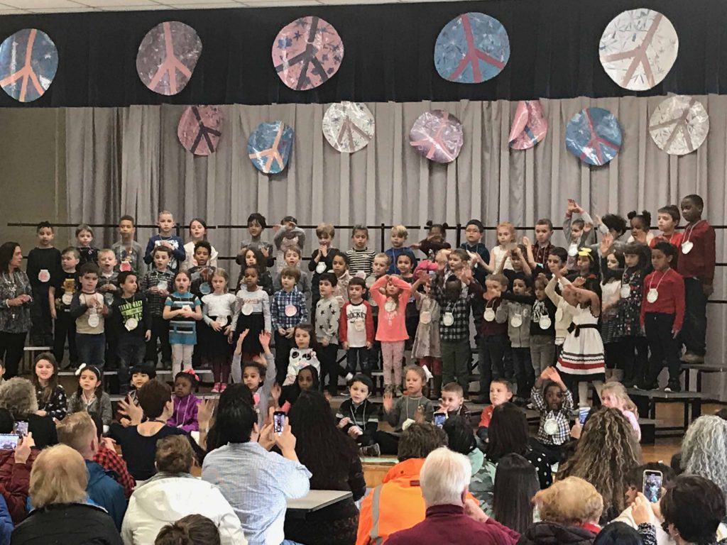 large group of kindergarten students on stage with parents in the audience, many videoing