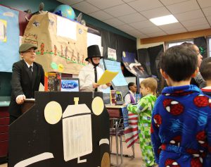 Fifth grade boy in a cap stands behind a cardboard car. Next to him is a boy dressed as Abraham Lincoln in top hat and beard talking to students.