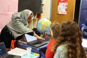Fifth-grade girl wearing a hijab points to a computer as she talks to two younger students. There is a globe on the table next to her