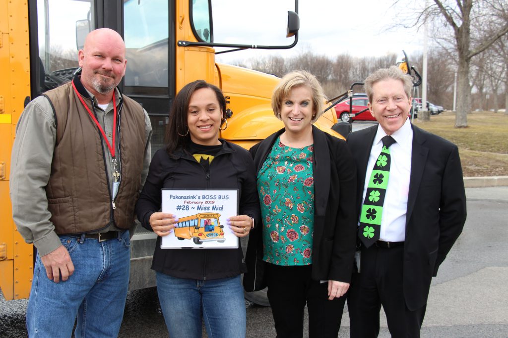 Four adults stand by a school bus. One is holding a certificate for being named Boss Bus for the month of February.