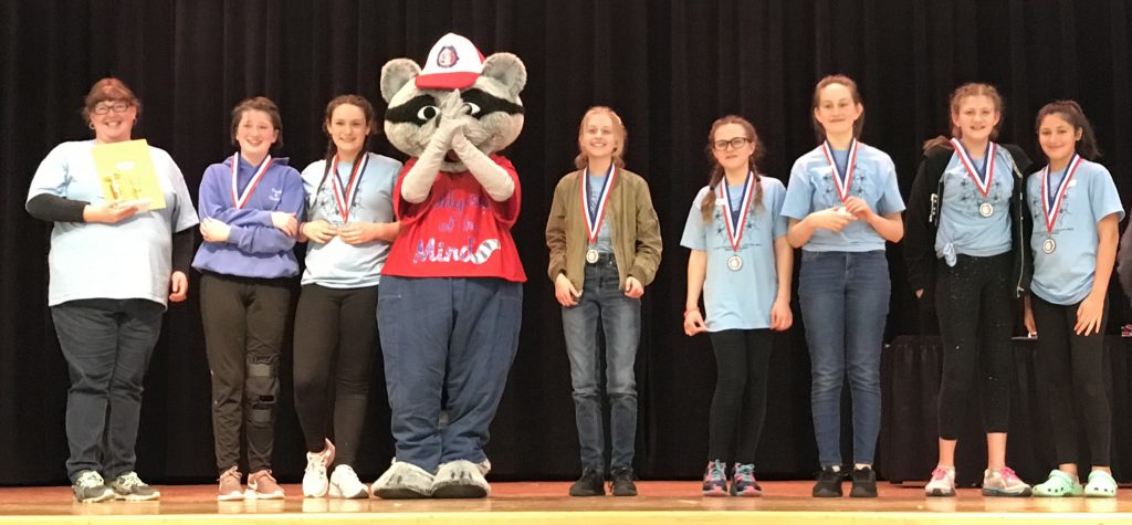 Seven middle school girls, dressed in light blue tshirts on a stage with their coach at one end and the Odyssey of the Mind mascot, a red-shirted raccoon.