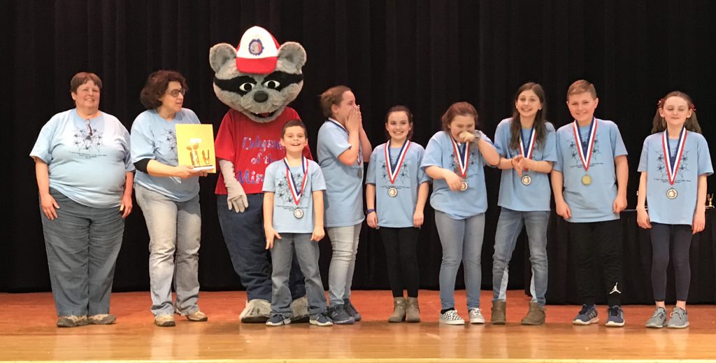 Seven elementary school students, two boys and five girls, wearing light blue tshirts with  metals around their necks standing on a stage with their two coaches at left and in the center the Odyssey mascot, a raccoon character dressed in a red tshirt