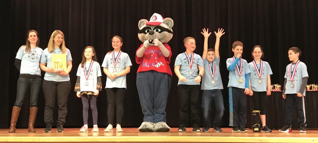 Seven elementary students smiling broadly - one with his arms straight up in the air - all wearing light blue tshirts. Their two coaches are at the left with the Odyssey mascot, a raccoon wearing a red tshirt.