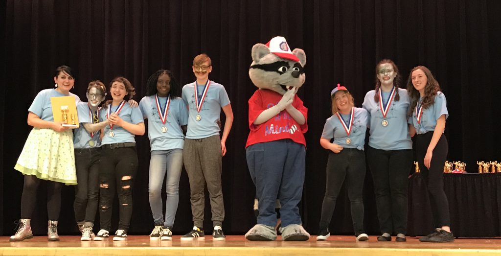 Seven high school students wearing light blue tshirts and first place medals smile and hug on stage with the Odyssey mascot