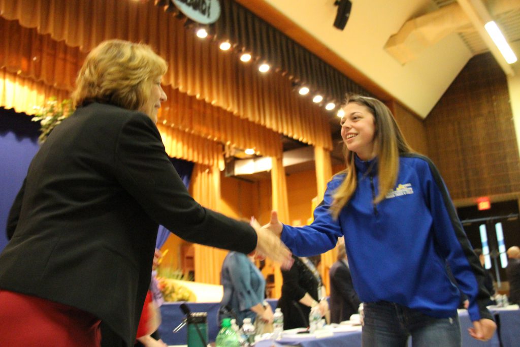 a young woman in blue jacket shakes hands with a blonde woman with stage in the background.