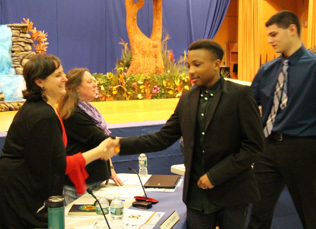 Board member in red blouse and black jacket smiles and shakes hands with male student athlete dressed in button-down shirt and jacket.