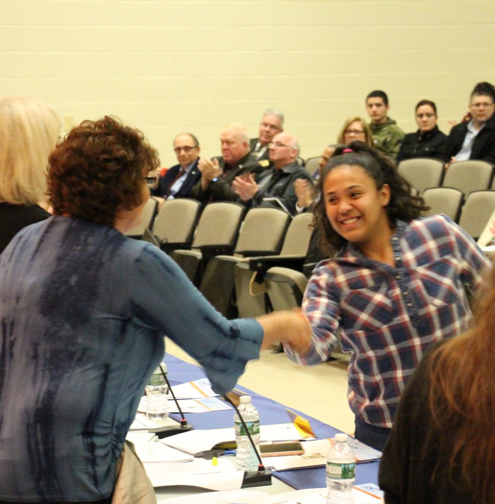 A high school girl with long dark hair dressed in a plaid shirt shakes hands with a board member in a blue shirt