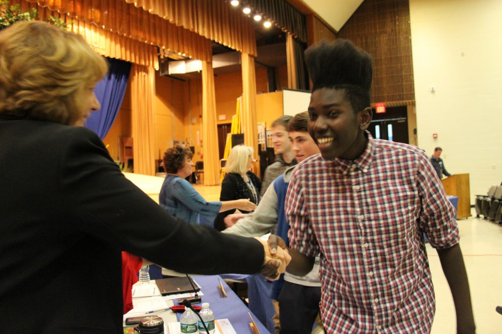 A male high school athlete smiles while shaking hands with the assistant superintendent.