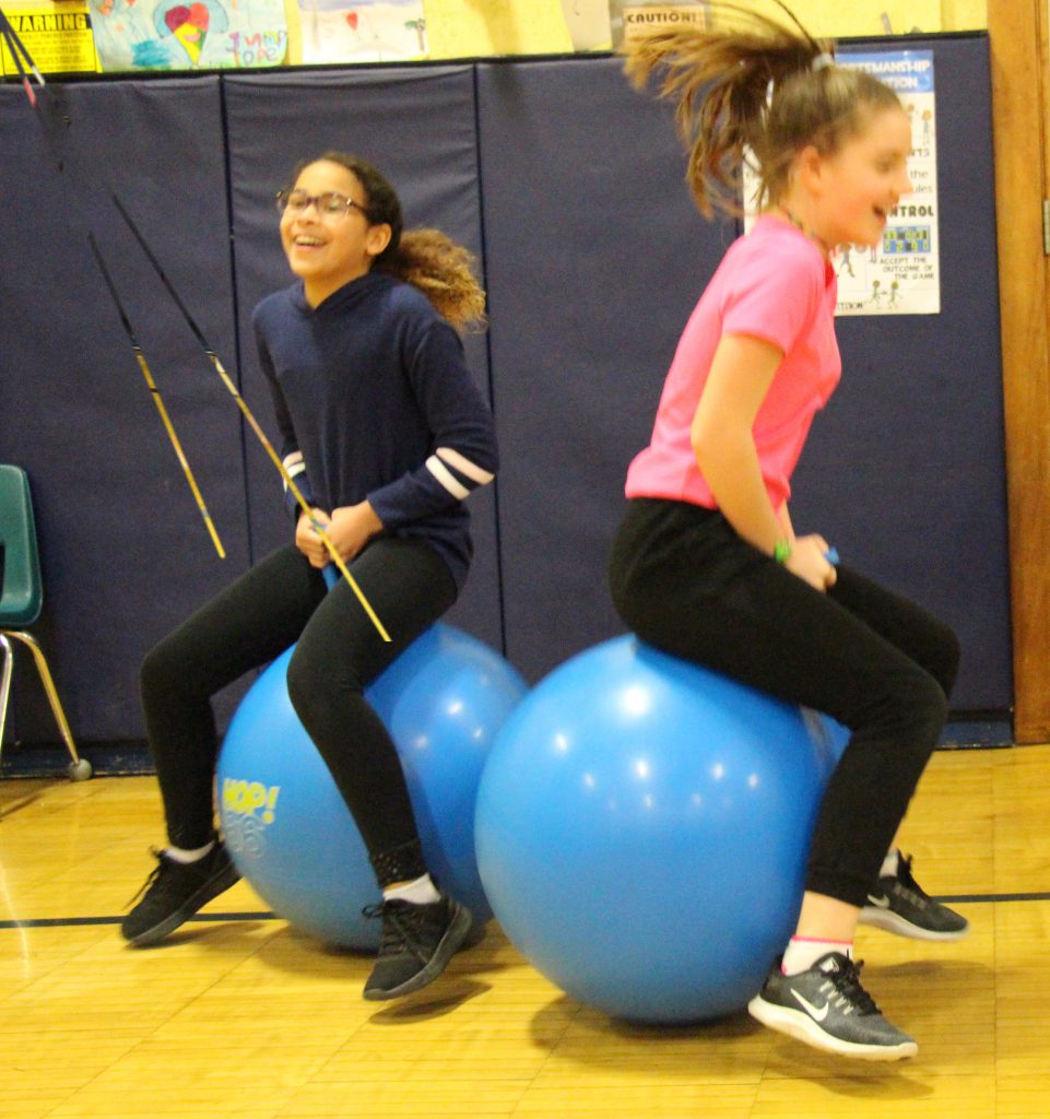 Two fifth-grade girls bounce on bright blue, large rubber bouncing balls.