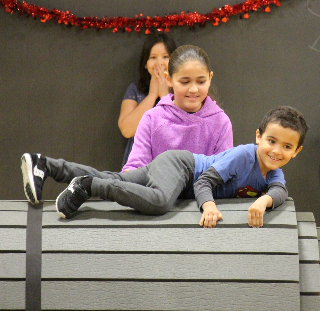 Small kindergarten boy takes part in an obstacle course, climbing over a large gray barrel-type structure while an older elementary student helps him.