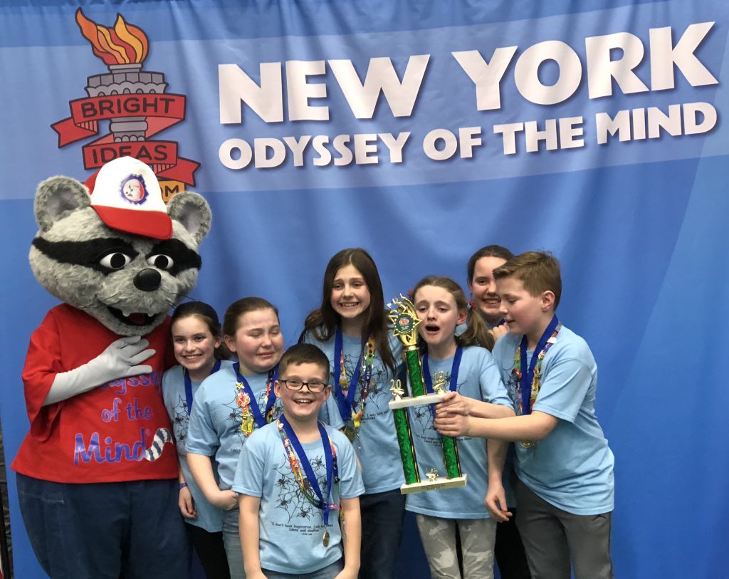 Seven elementary students have medals around their necks and hold their trophy with the Odyssey of the Mond mascot, a large raccoon character dressed in red.