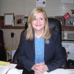 Woman with shoulder-length blonde hair smiling while sitting at her desk. Wearing a blue jacket and blouse.
