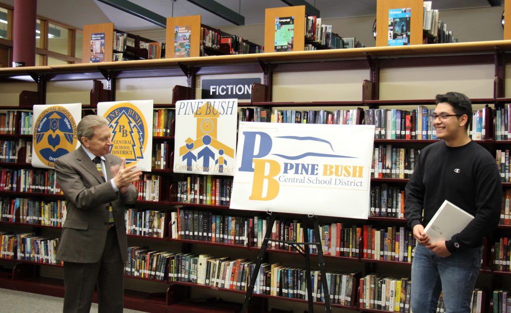 A man on the left claps while a young man on the right smiles as they both look at a poster-size rendering of the new Pine Bush logo. 