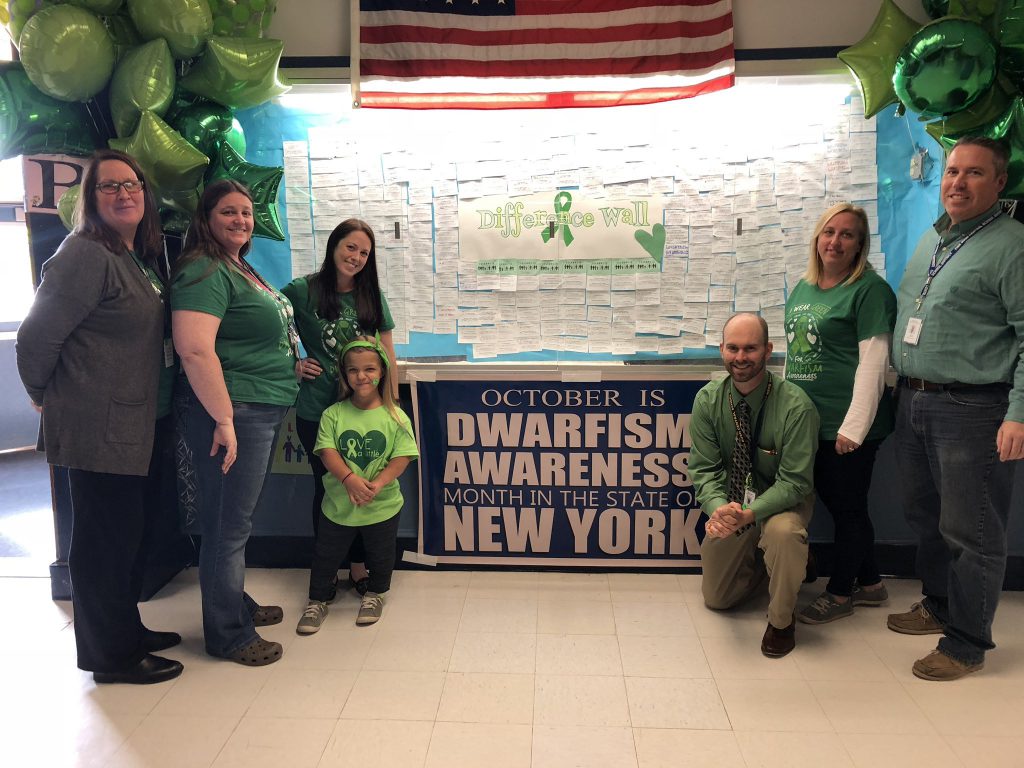 Six adults and one middle school student dressed in green stand in front of a sign for the difference wall to mark dwarfism awareness.