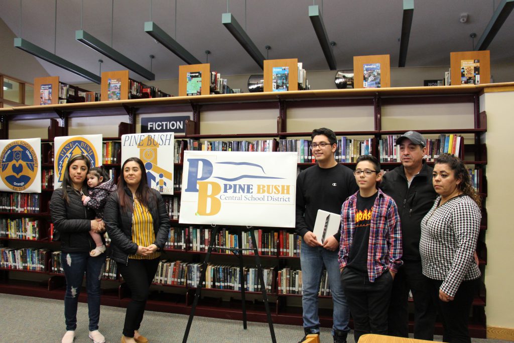 A large rendering of the new Pine Bush logo is in the center with two women on the left, one holding a baby, two men, a boy and a woman on the right. In the background are bookshelves filled with library books.