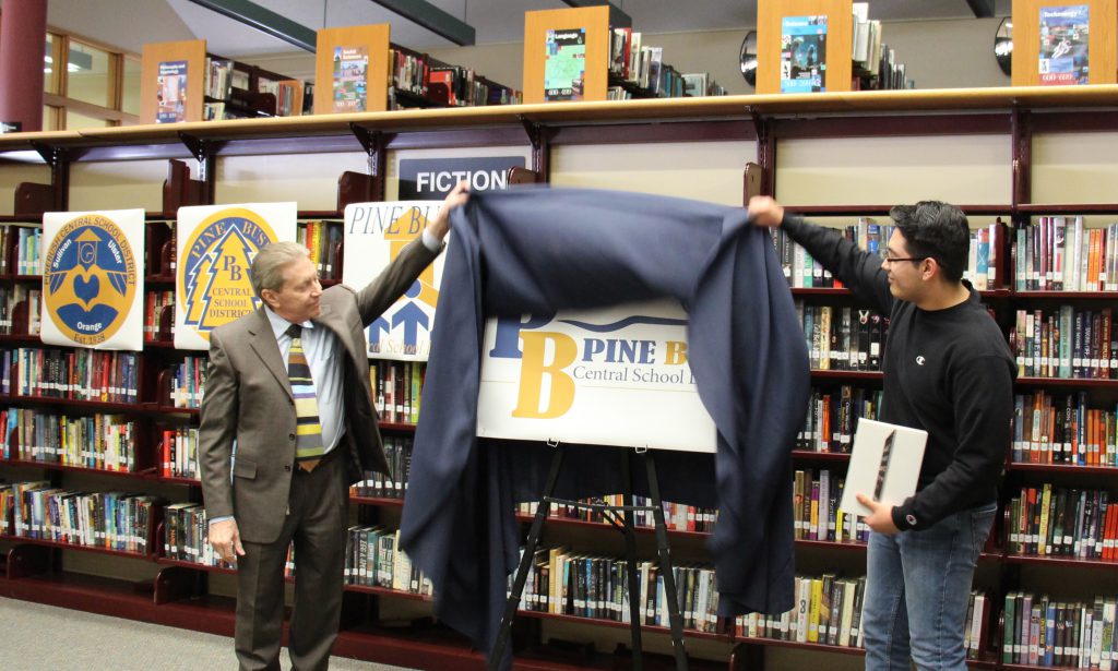 Men on their side of a poster lift off a blue curtain showing a poster of the new Pine Bush logo.