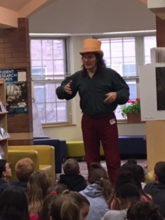 Students sitting on the floor listening as a may wearing a tan top hat acts out a story