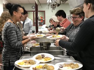 In the kitchen, helpers are putting plates of food on large trays