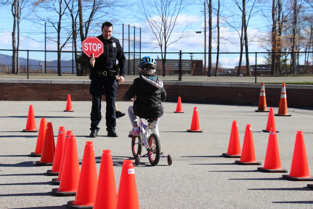 Kindergarten student on a bike with training wheels riding between cones with a police officer in front holding a stop sign.