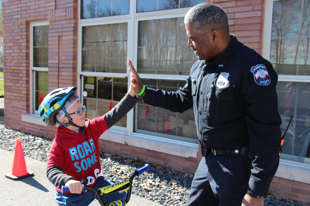 Police officer high fives a kindergarten student on a bike wearing a helmet and glasses smiling.