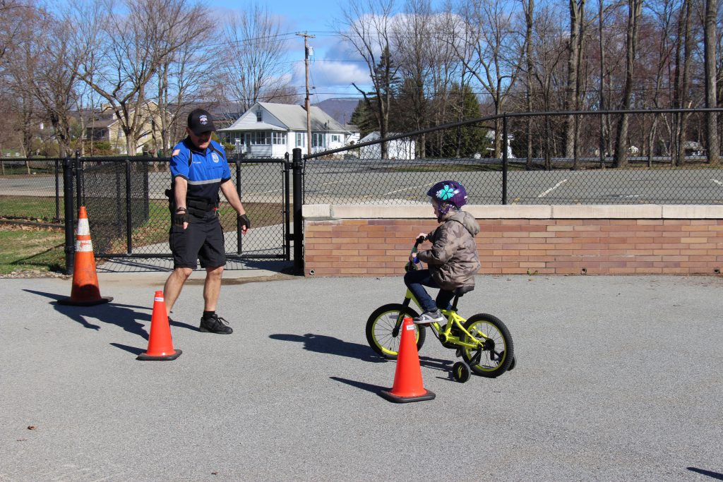 Officer in shorts and short-sleeve shirt and a hat points to a place between orange cones while kindergarten student rides her bike through it