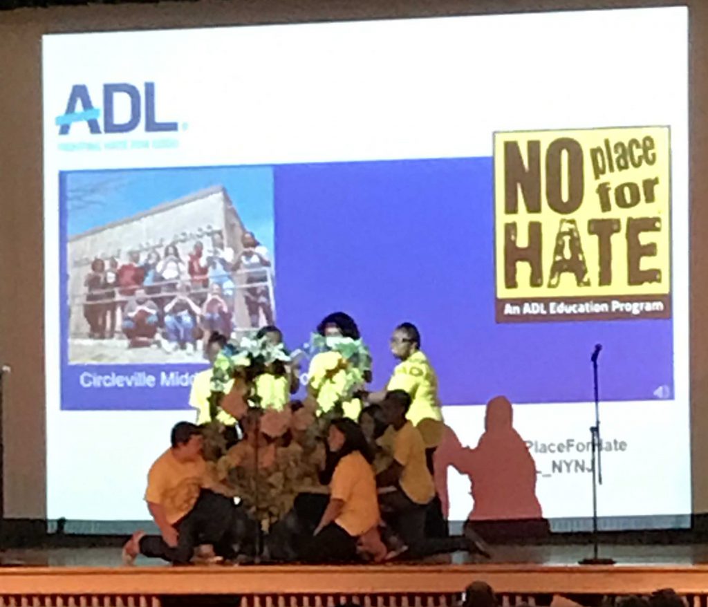 A group of students in yellow No Place For Hate tshirts are grouped on the stage, holding flowers in the shape of a heart. They are in front of a large screen with the graphic No Place For Hate