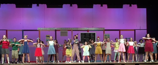 Several members of the cast of Junie B. Jones dressed in costume sing in front of a pink and purple background