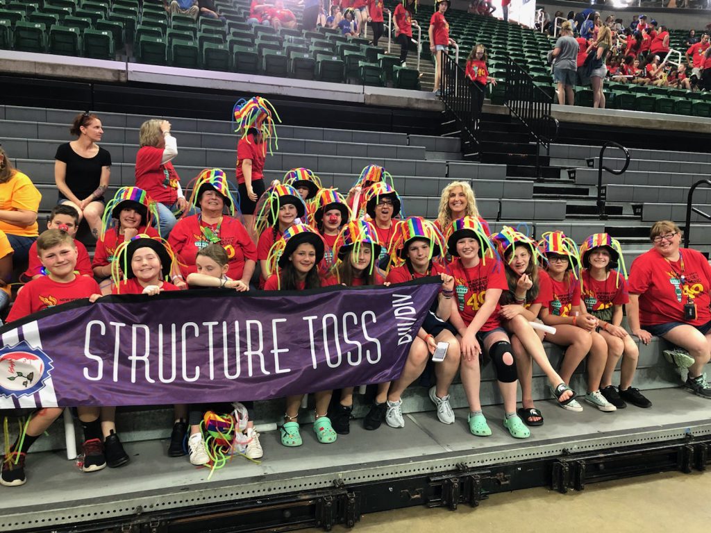 About 15 students and several adults all wearing red shirts holding a banner with their Odyssey of the Mind problem name, Structure Toss.