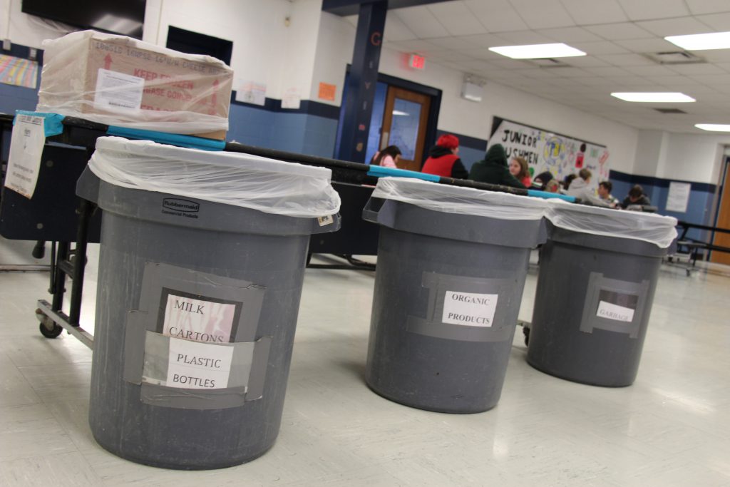 Three gray trash cans marked with small white signs showing which one is for recycling, trash and organic material