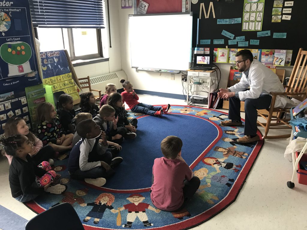 Man in a white doctor's coat sits and talks to young students who are sitting on an oblong rug.