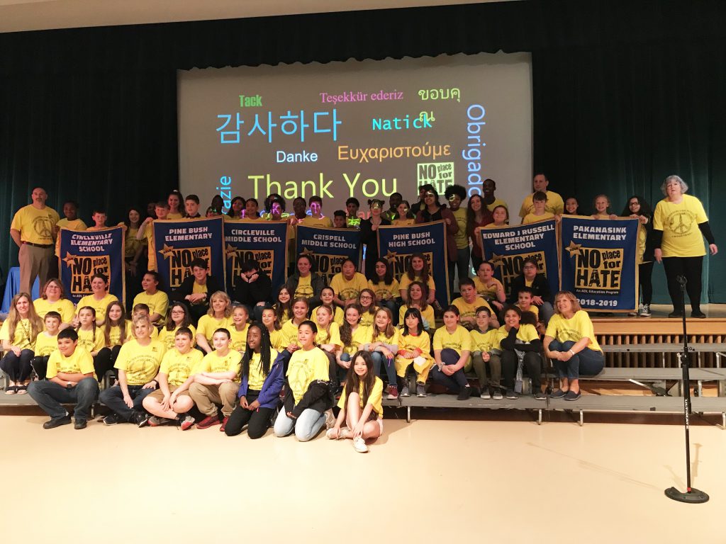 A group of 100, mostly students, sit on the steps of the stage. All are wearing No Place For Hate tshirts. Screen in the back has Thank YOu in many languages