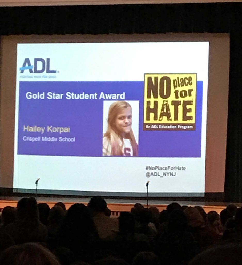 A large screen with the blue and gold graphics of the ADL, showing No Place For Hate logo and a picture of Crispell student Hailey Korpei.