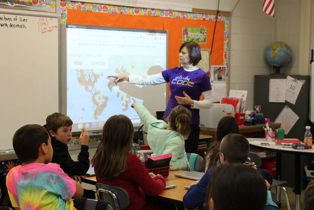 Teacher in purple t shirt that says "Girls Who Code" points to a screen showing a map of the world indicating all the places the day of code is happening. Students are pointing too.