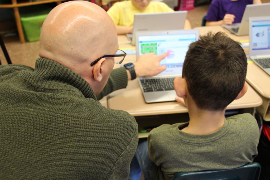 A man in a green sweater points to a chromebook screen while instructing a student sitting next to him