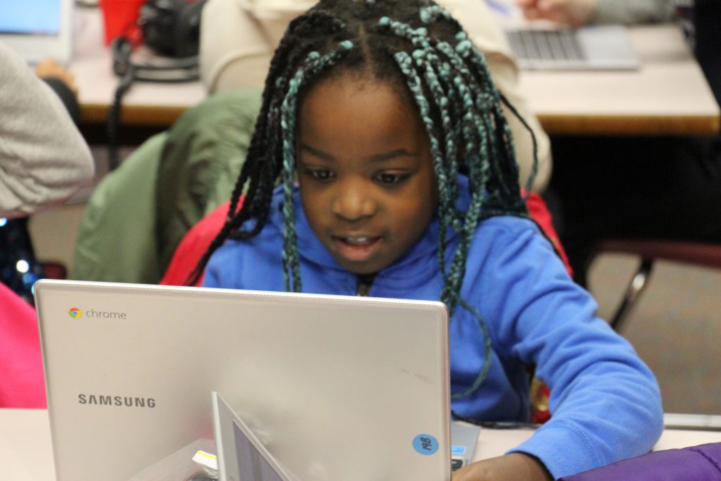 Young girl with a purple sweatshirt and long braids watches her chrome book screen intently