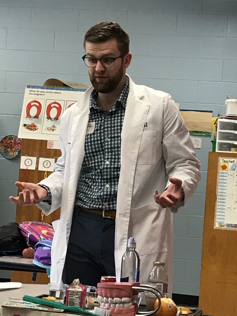 Man in a white doctor's coat, dark hair and glasses talks while on the table in front of him are dental related objects.