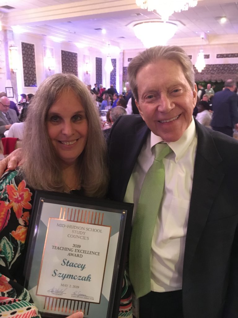 A woman with shoulder length hair and flowered dress smiles and holds a certificate. Next to her with his arm around her is a smiling man with suit and green tie.