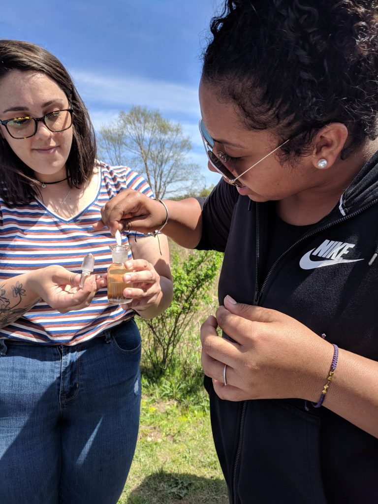 One student holding a samle of pond water in a small container while another student uses a dropper to take water out.