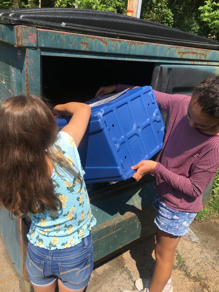 Two students dump the contents of a blue recycle bin into a larger recycle dumpster.