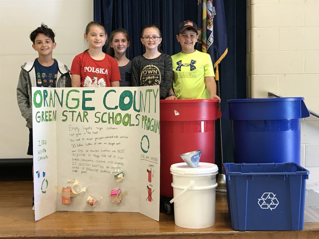 Five fourth-grade students stand behind a poster board and next to a red trash can, blue recycle bins and a white container with a funnel.
