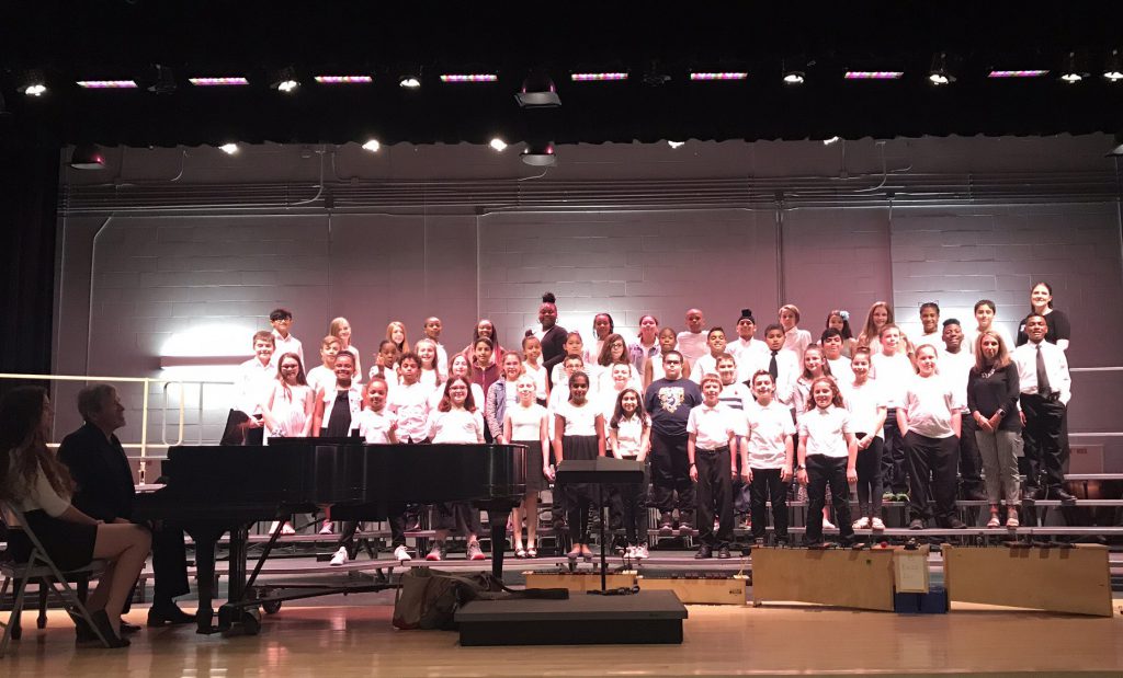 More than 50 fifth grade students dressed in white shirts and black pants stand on stage ready to perform. Adults flank them.