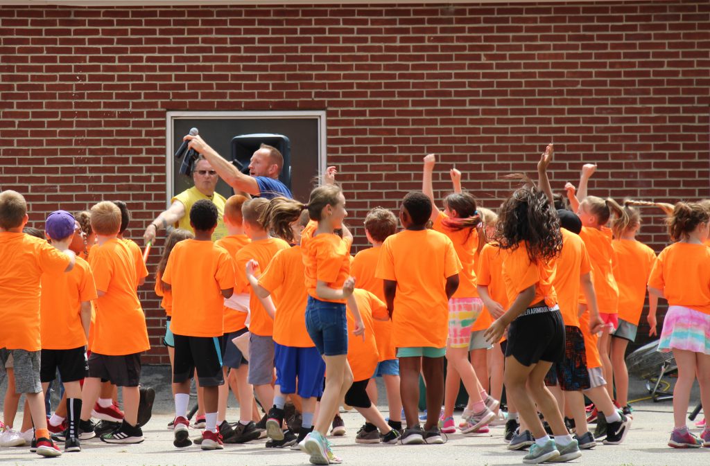 A large group of elementary students, all wearing orange t-shirts, dancing