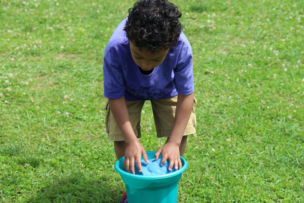 Little boy in purple shirt and tan shorts leaning over a bucket of water with a sponge in his hands.