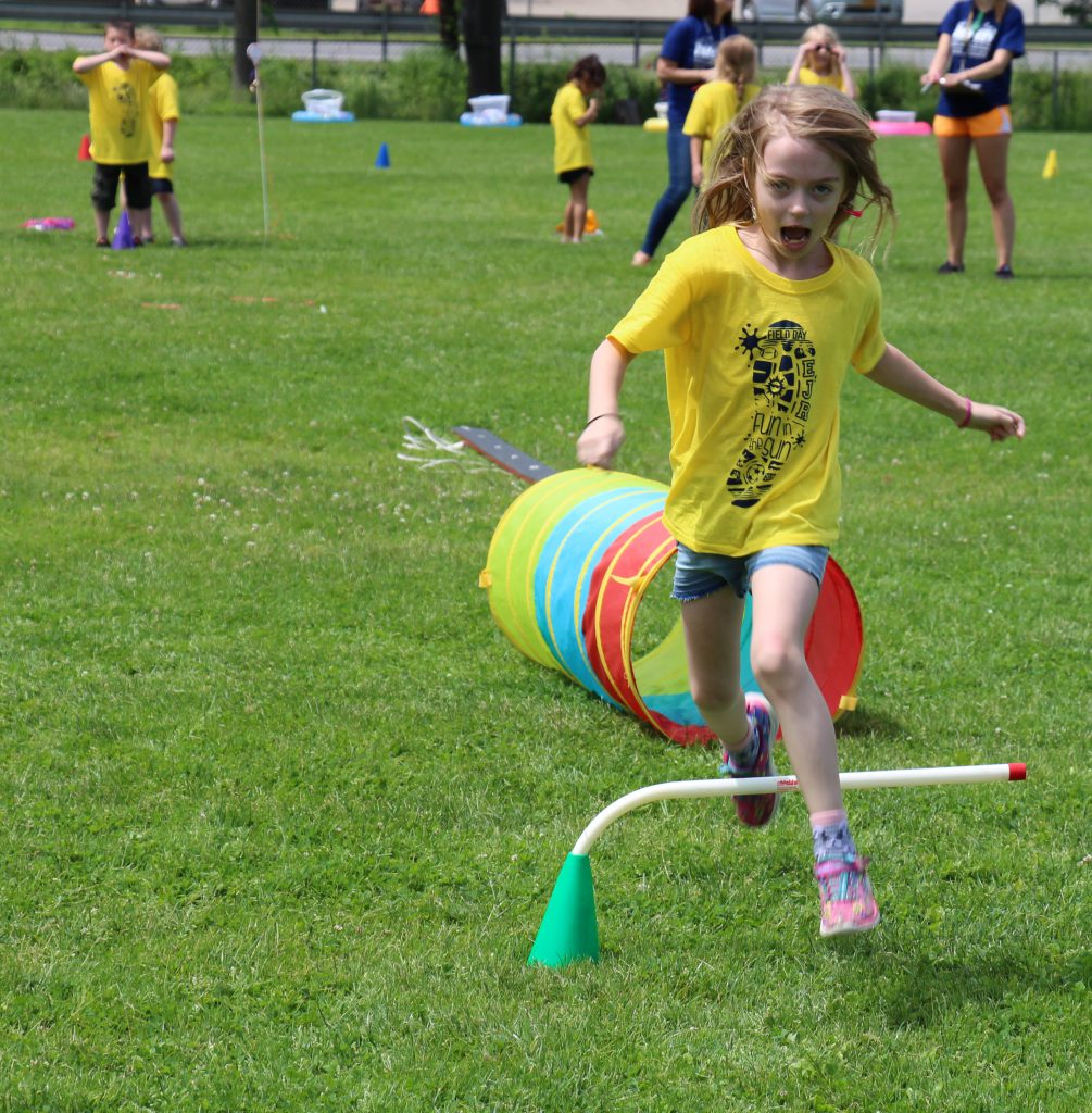 Elementary school girl wearing a yellow t-shirt jumps over a bar on the obstacle course.