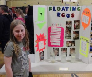 Girl with long hair wearing a gray shirt stands in front of her tri-fold board with an experiment about floating eggs