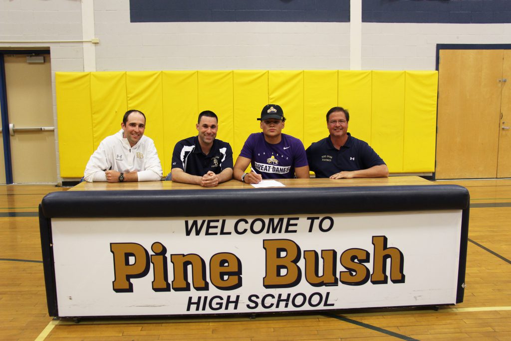 Four men at a table that says Welcome to Pine Bush High School. Third from left is wearing a hat and a U Albany t shirt.