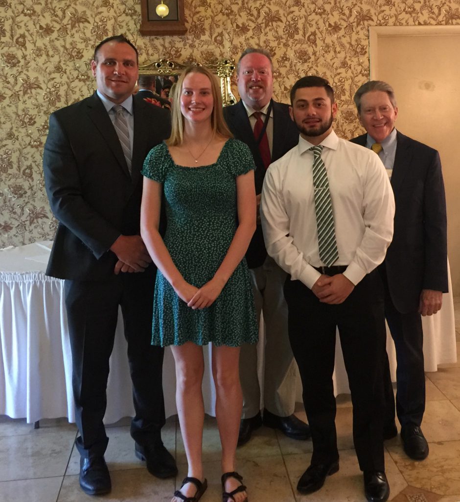 Three men in suites stand behind two high school senior athletes, one in a green dress, the other in a white shirt and tie.