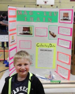 A young elementary school boy stands in front of his tri-fold board showing his science project.