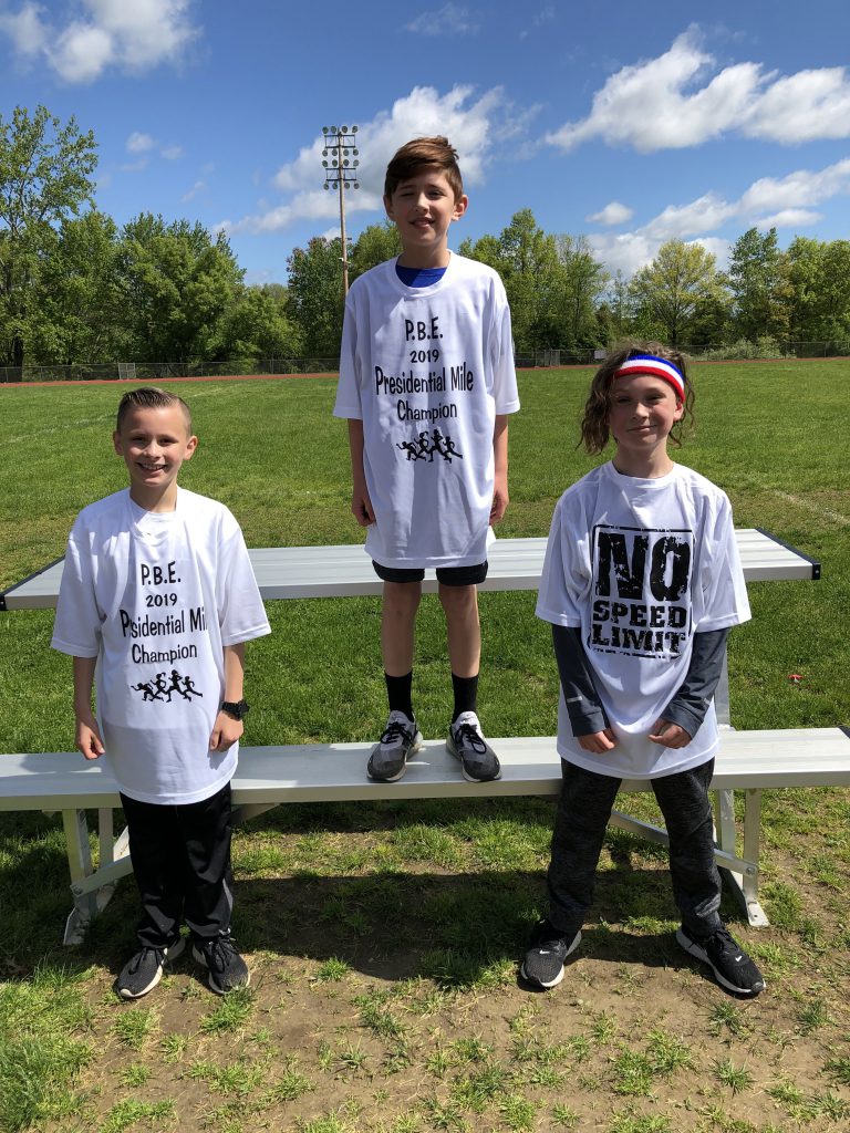 Three fourth-grade boys stand wearing their white presidential mile t shirts. First place winner in center stands higher.