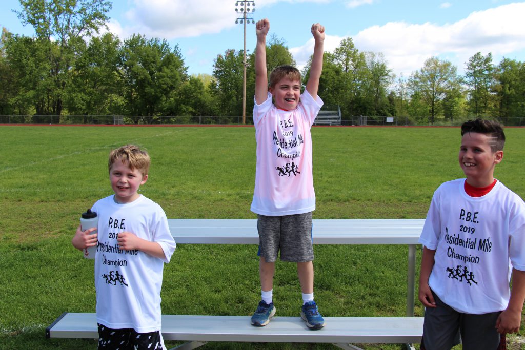 Third-grade boys top three finishers wearing white presidential mile t shirts. The first place winner in center has his arms straight up over his head.